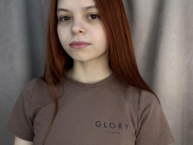 Sadie Yankovitz a de longs cheveux roux, une peau pâle, des yeux marrons, et elle porte une chemise marron avec l'inscription "GLORY MILANO". Elle regarde l'appareil photo avec un léger sourire..