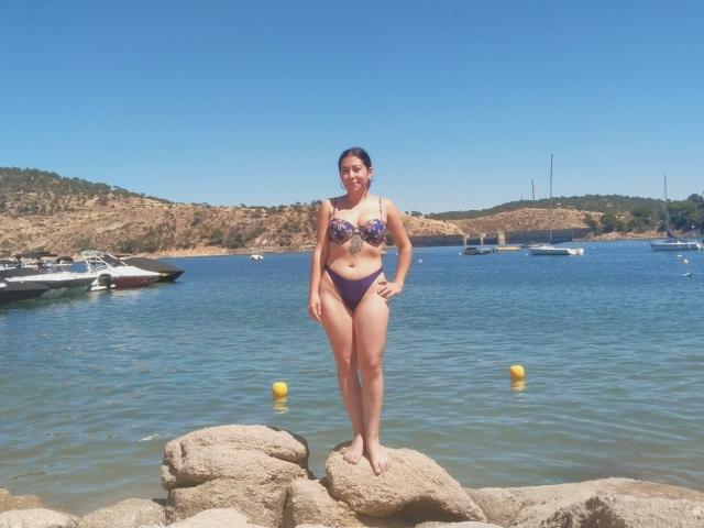 AmaraParker stands on rocks in a blue bikini, showing her pussy-sized tits in a purple bikini top and matching bottom, with a clear blue sky and water behind.