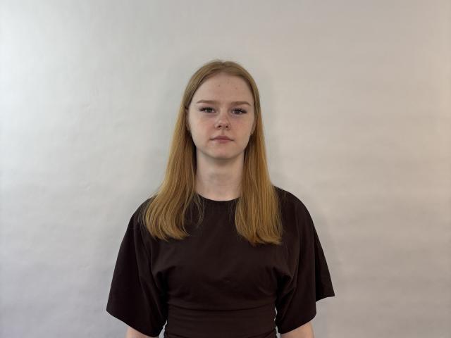 Shakia Ivester, met recht, halflang blond haar, draagt een donkerbruin shirt met korte mouwen en staat tegen een effen witte muur, waarbij ze iets naar links kijkt met een neutrale uitdrukking..