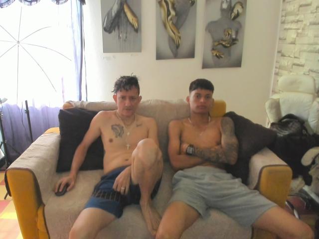 CristianAndLuke, sem camisa e com tatuagens, estão sentados em um sofá bege. Cristian tem uma tatuagem no peito e uma lesão no joelho, vestindo shorts azuis. Luke tem tatuagens no braço e está usando shorts cinzas..