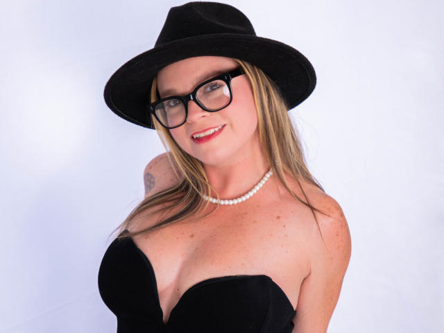 PamelaColin with black hat, glasses, pearl necklace, and black strapless dress showing her big tits, smiling, blonde hair, and tattoo on right shoulder.