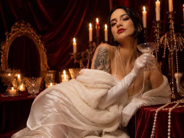 Celeste Ocampo, avec des cheveux noirs, du rouge à lèvres, une robe en satin blanc, des gants blancs, tenant un verre de martini, une étole en fourrure blanche, un tatouage sur le bras droit, entourée de bougies et de perles..