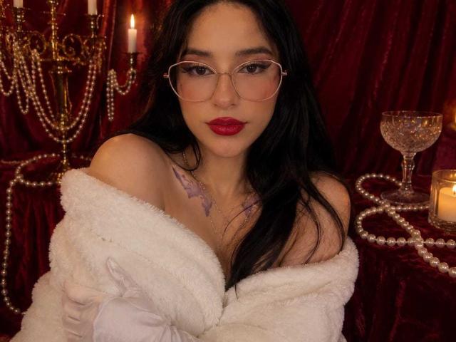 Celeste Ocampo, avec des lunettes, du rouge à lèvres rouge, un manteau blanc et duveteux porté à une épaule, dévoilant sa poitrine avec des tatouages violets, cheveux foncés, fond en velours rouge, bougies, verre et collier de perles..