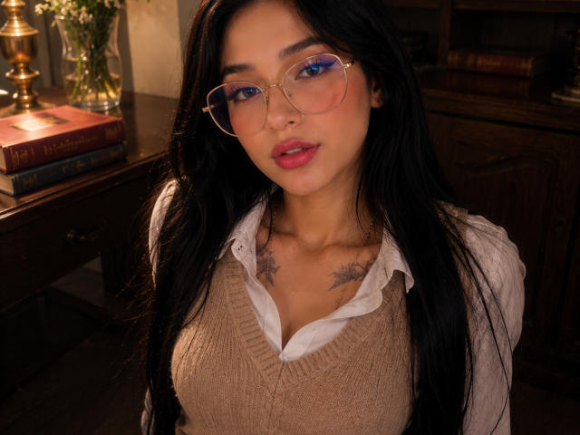 Celeste Ocampo, avec de longs cheveux noirs, des lunettes, des lèvres roses, un pull beige, une chemise blanche, de petits tatouages sur la poitrine, un fond en bois marron, une bibliothèque et une lampe. Ses seins sont visibles sous le pull..