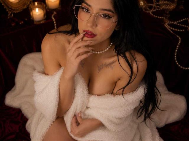 Celeste Ocampo mit langem, schwarzem Haar, Brille und einer Perlenkette, die ihre Brüste mit einem weißen, flauschigen Bademantel bedeckt, ein Finger in der Nähe ihrer roten Lippen, Kerzen im Hintergrund..