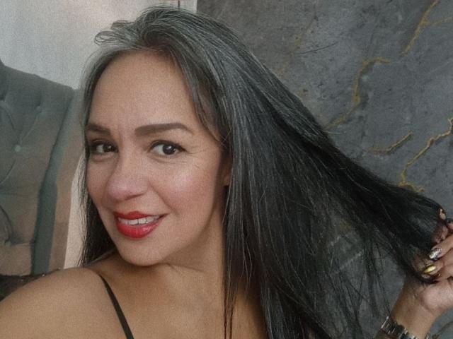 LailaCastillo has long black hair, smiling with red lipstick, holding a lock of her hair, showing her dark eyes, and wearing a black spaghetti strap top.