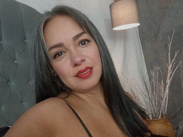 LailaCastillo has long black hair, red lipstick, and brown eyes. She's wearing a black spaghetti strap top, smiling with her boobs partially visible. She's in a bedroom.