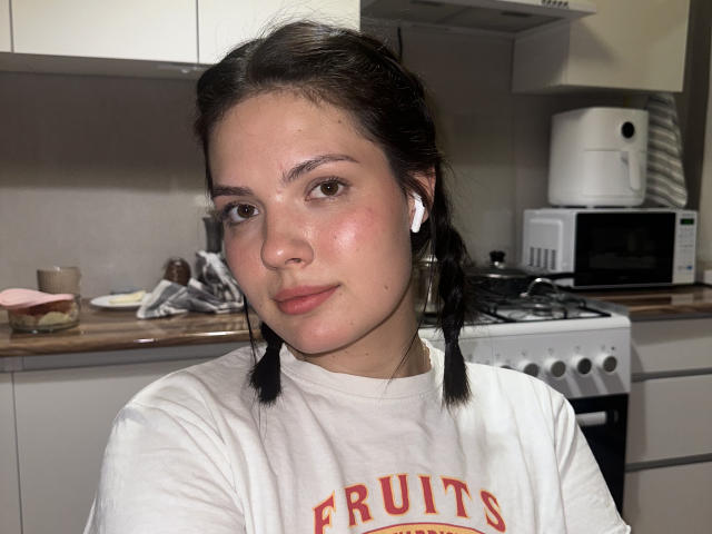 HortenseSpruit with puffy cheeks, wearing white earbuds, has braided black hair, and is dressed in a white shirt with red text. She's in a kitchen with appliances.