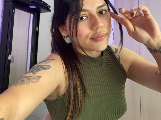 SophiaMarx with long brown hair, green ribbed top, tattoos on arms, smiling, making a peace sign, brown eyes, light skin, small nose, slightly flushed cheeks, pink lips.