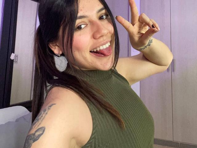 SophiaMarx with pigtails, green sleeveless ribbed top, tongue out, making a peace sign, tattoo on upper arm, light skin, black hair, smiling, visible boob, and small ass.