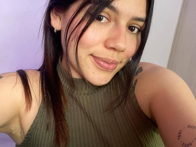SophiaMarx has a cute smile, small tits, and tattoos on her arms. She's wearing a green ribbed sleeveless top, with black hair and subtle makeup.