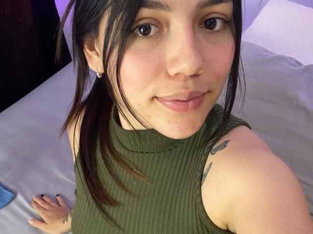 SophiaMarx with green ribbed sleeveless top, brown pigtails, nose ring, small tattoos on right arm, subtle smile, light skin, and purple-lit bedroom, holding a grey bedsheet.