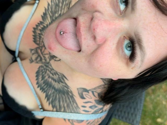Prettyjosi-hot with tongue out, black bra, tattoos on chest and shoulders, blue eyes, pierced tongue, short brown hair, fair skin, visible cleavage.