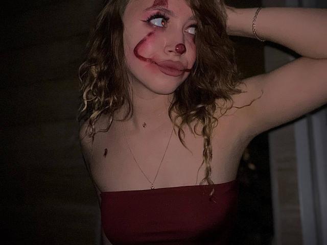 Alane Gaudenzi, con maquillaje que simula sangre, lápiz labial rojo y un piercing en la nariz, lleva un top rojo sin tirantes, tiene el cabello rubio rizado, un brazo levantado y una pulsera, en una habitación oscura..