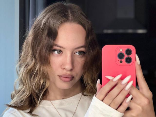 Alane Gaudenzi, met golvend bruin haar, houdt een roze iPhone vast, kijkt naar de camera, draagt een witte trui, heeft lange witte nagels en een neutrale uitdrukking..