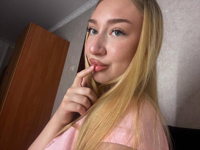 Joette Shigeta, met blond haar, blauwe ogen en roze lippen, die haar vinger op haar mond zet, draagt een roze geribbelde top en kijkt naar de camera..
