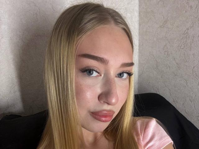 JoetteShigeta has blonde hair, blue eyes, and pink lips. She's looking at the camera with a slight smile, wearing a pink top, and has long, straight eyebrows.