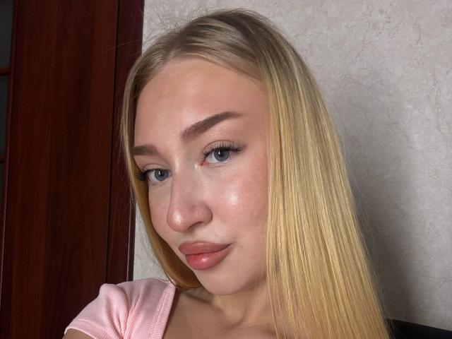 Joette Shigeta a les cheveux blonds, les yeux bleus et les lèvres roses légèrement retroussées. Elle porte un haut rose, et son visage est légèrement brillant avec du maquillage visible..
