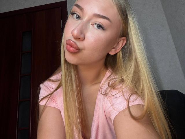 Blonde JoetteShigeta met volle, roze lippen, draagt een roze top en heeft lang, steil haar, lichte huid en groene ogen, op een selfie. Haar borsten zijn licht zichtbaar..