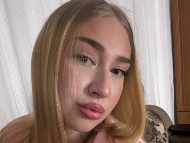 Joette Shigeta tem cabelo loiro, lábios rosados e olhos azuis com sobrancelhas bem definidas. Ela está olhando para a câmera com um leve sorriso, e sua mão está perto do rosto..
