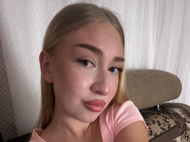 JoetteShigeta with blonde hair, blue eyes, and pink lips, wearing a pink shirt, looking at the camera with a slight pout, eyebrows shaped, subtle makeup, in a selfie.