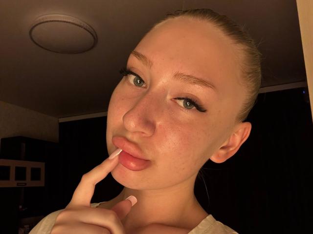 JoetteShigeta with pale skin, blonde hair in a bun, green eyes, and freckles, sucking her index finger, wearing a white top, in a dimly lit room.