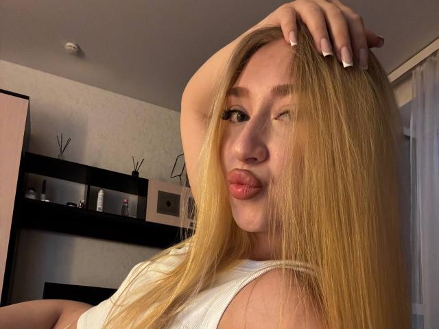 Joette Shigeta met blond haar, knippend, met een ondeugende mond, hand in haar haar, draagt een witte top, toont haar achterwerk op de achtergrond, lange nagels, schattig gezicht, zachte belichting, binnenomgeving..