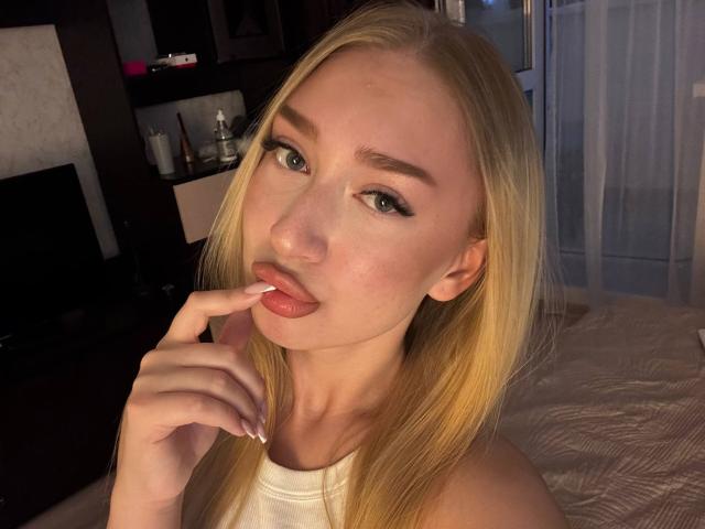Blonde JoetteShigeta with green eyes, white tank top, and finger in mouth, showing her glossy pink lips, with her hand near her face in a dimly lit room.
