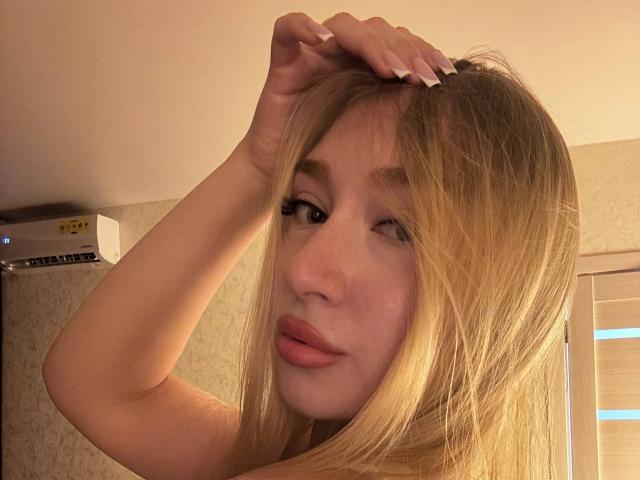 Joette Shigeta met blond haar, knippend, hand in haar, roze lippen, lichte huid en lange nagels, die haar schattige gezicht en lichte glimlach laat zien. Airconditioning in de achtergrond..