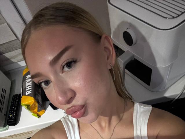 Joette Shigeta heeft blond haar, blauwe ogen en volle, roze lippen, draagt een witte tanktop, heeft een kleine neus en een lichte huid, en een zilveren ketting..