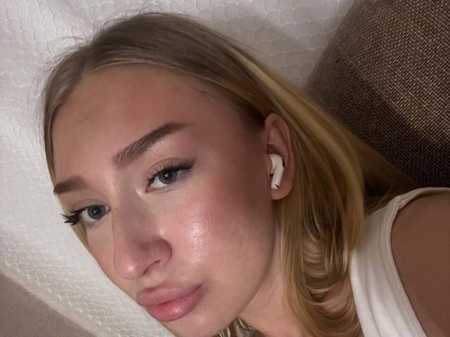 Joette Shigeta heeft blond haar, blauwe ogen en witte oordopjes, met een glanzende, lichtroze huid en subtiele make-up, en kijkt iets opzij. Ze draagt een wit topje zonder mouwen..