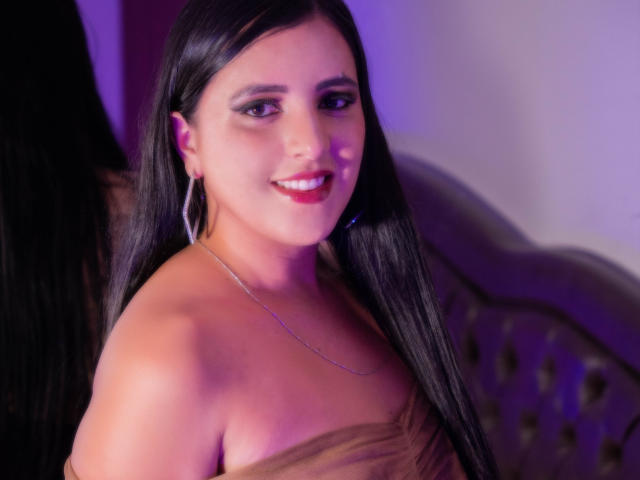 AlanaSantos has long black hair, red lipstick, and a smile, wearing a strapless brown dress, hoop earrings, and a silver necklace, with purple lighting in the background.
