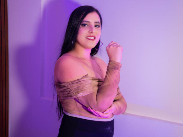 AlanaSantos with long black hair, brown off-shoulder top, black pants, purple light, smiling, arms crossed, small hoop earrings, fair skin, cute, sexy, standing against white wall.