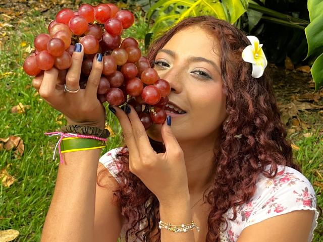 Mia Bianchi, met krullend rood haar, een witte bloem in haar haar, blauwe nagellak, die rode druiven eet, een bloemenprint top draagt, een gele armband en een roze lint om haar pols..