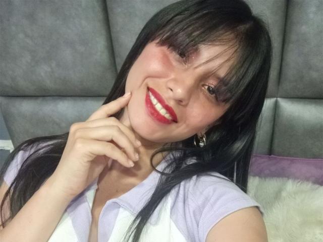 MadissonFontaine ha lunghi capelli neri, rossetto rosso ed è sorridente, con un dito che tocca la guancia. Indossa una camicia bianca e viola, che mette in mostra il suo seno..