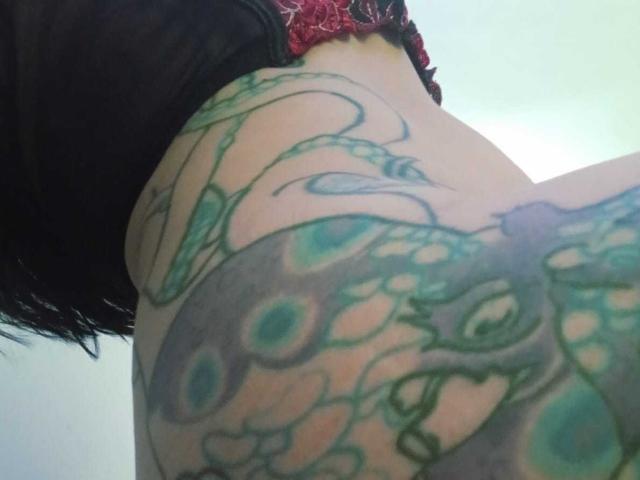 ZarayFeet's tattooed thigh, featuring a green peacock design with blue eyes, partially visible black top, and red lace. Upper body angle, focusing on the tattoo.