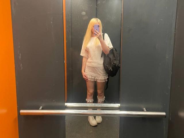 Lorine Hamway, avec de longs cheveux blonds, portant une chemise blanche à manches courtes et une mini-jupe ornée de dentelle, tenant un sac noir, debout devant un miroir, en train de prendre un selfie..