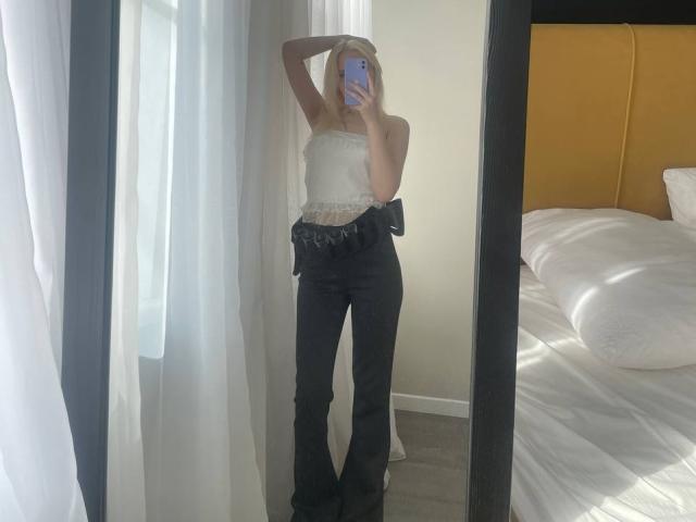 Lorine Hamway con i seni scoperti, pantaloni neri, top di pizzo bianco, mano tra i capelli, selfie allo specchio, che tiene il telefono, in piedi vicino al letto con testiera gialla, tende bianche, luce solare..