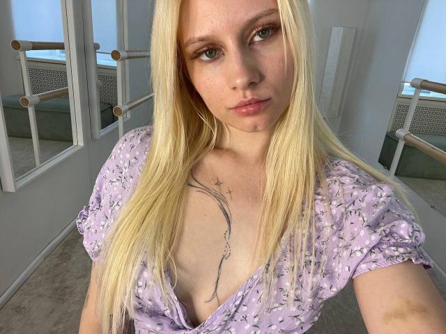 Lorine Hamway a de longs cheveux blonds, des yeux verts et un tatouage sur la poitrine. Elle porte une robe fleurie violette qui dévoile ses seins, dans une pièce lumineuse..