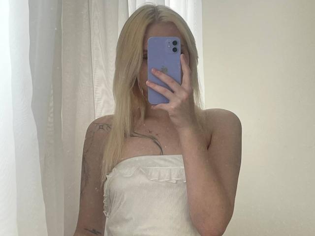 Lorine Hamway, bionda, con un abito bianco senza spalline, tiene in mano un telefono viola, si copre il viso, mostrando il seno, ha un tatuaggio sul braccio sinistro, e sullo sfondo si vede una tenda bianca..
