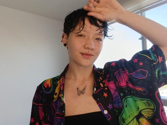 LissyLuxe with short black wet hair, wearing a colorful star and mushroom print shirt, black top, butterfly tattoo on chest, black ear plugs, smirking, right hand in hair.