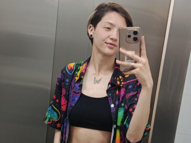 LissyLuxe with short brown hair, wearing a colorful shirt over a black sports bra, holding a phone, showing a small butterfly tattoo on her chest, smiling in a mirror.