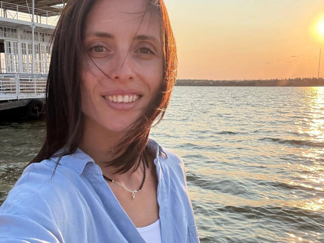 MilediElla - een aantrekkelijke vrouw met bruin haar, een blauw shirt, een wit T-shirt, een ketting, die lacht bij zonsondergang, met een weerspiegeling in het water, en een witte steiger op de achtergrond..