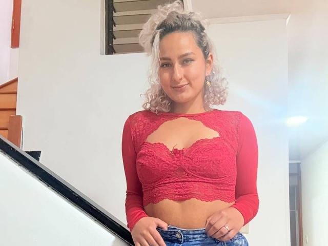 ElenaSaenz with curly pinkish hair, wearing a red lace crop top with cutout, showing her small tits, and blue jeans, standing near a staircase, smiling slightly.
