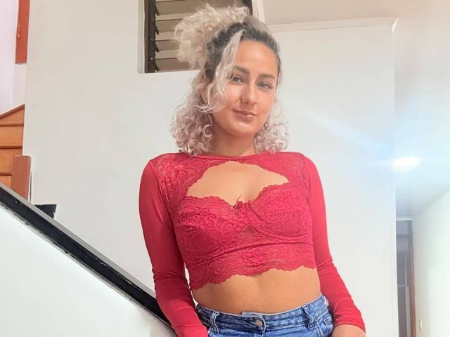 ElenaSaenz with curly blonde hair, wearing a red lace crop top with a cutout, showing her tits, and blue jeans, standing in a white-walled room.