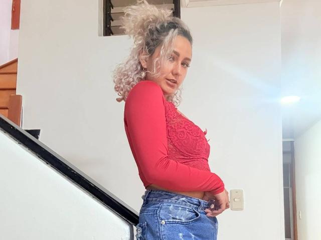 ElenaSaenz with curly blonde hair in a pink lace top and blue jeans, standing sideways, showing her tits, near a staircase. She's looking at the camera.