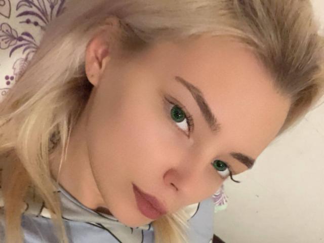 AdrieneDegennaro has blonde hair, green eyes, and is looking sideways with a slight smile. She's wearing a light blue shirt, and her makeup is subtle.