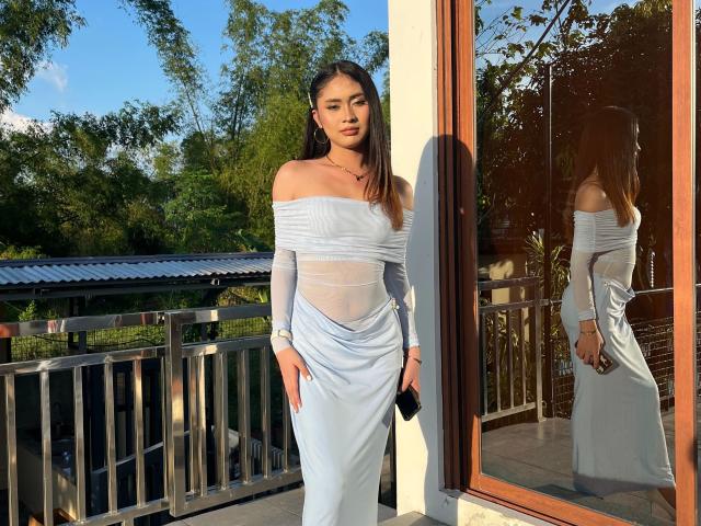 Sexygoddessjade-hot staat in het zonlicht, gekleed in een witte jurk met een open schouderlijn en een doorschijnende taille, en houdt een zwarte telefoon vast. Haar reflectie is te zien in het raam..