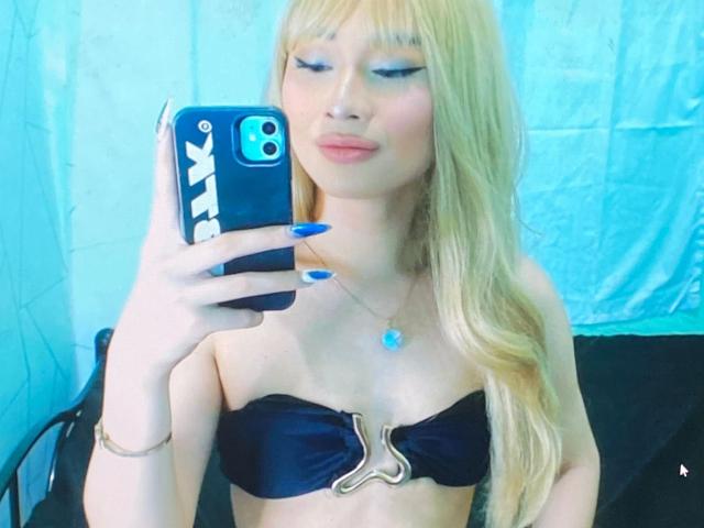 Sexygoddessjade-atractiva, con cabello rubio, uñas azules, top negro sin tirantes con cremallera dorada, sosteniendo un teléfono, fondo azul, mirando el espejo, pequeños senos, linda, labios ligeramente fruncidos, collar..