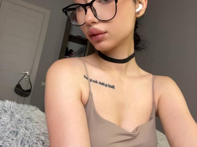 GlitchDoll with glasses, wearing a brown tank top, black choker, and earbuds, showing a tattoo on her chest. Her small tits and nose ring are visible.